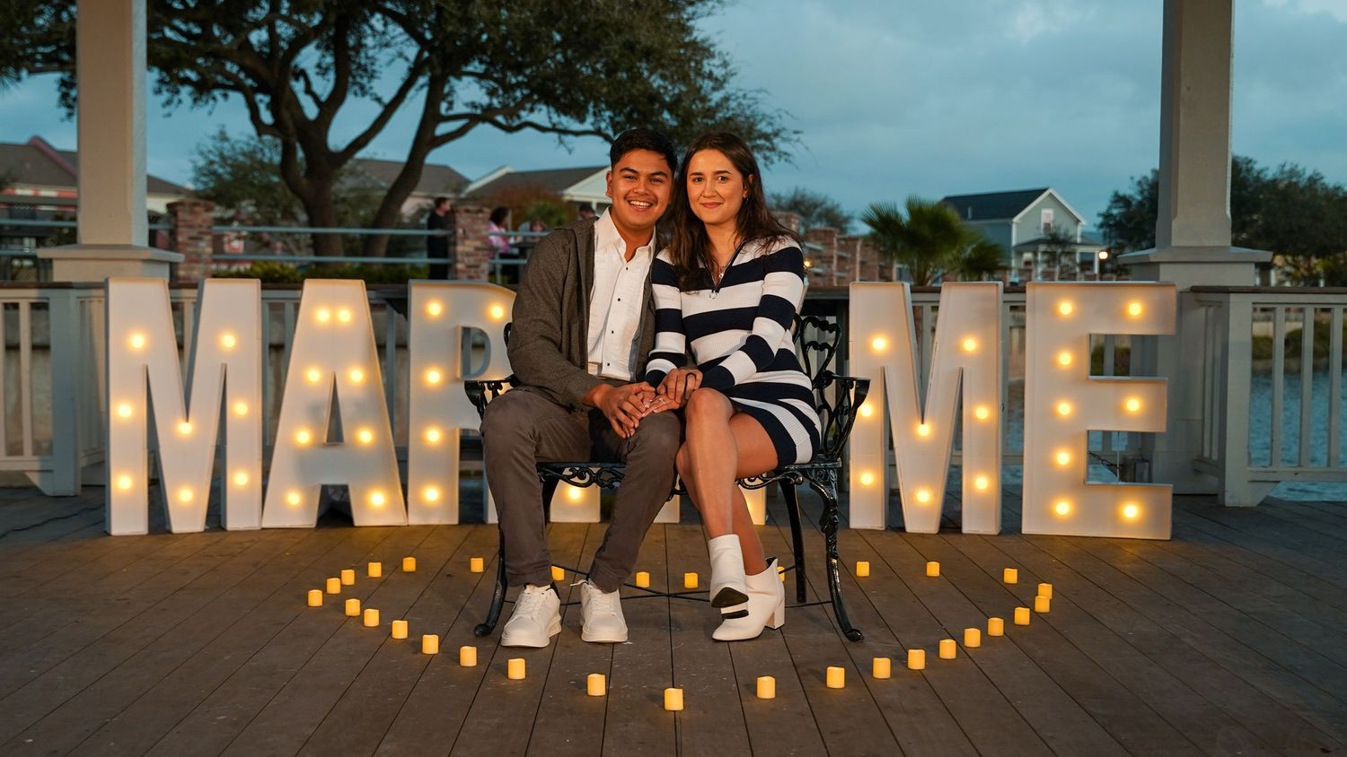 Couple seated in front of illuminated MARRY ME sign with candles at twilight.