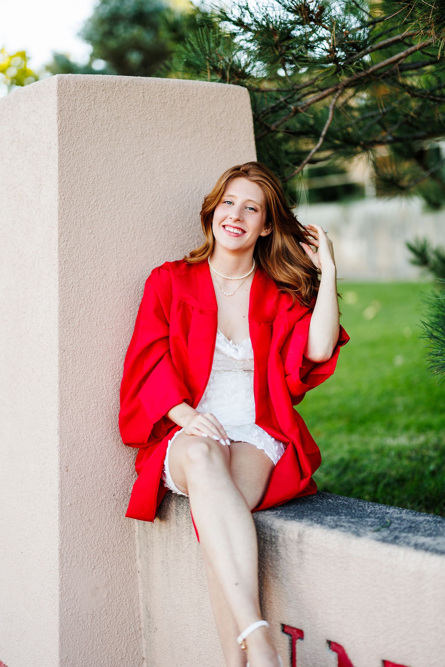 A stylish woman in a red blazer sits on a concrete ledge near evergreen trees.