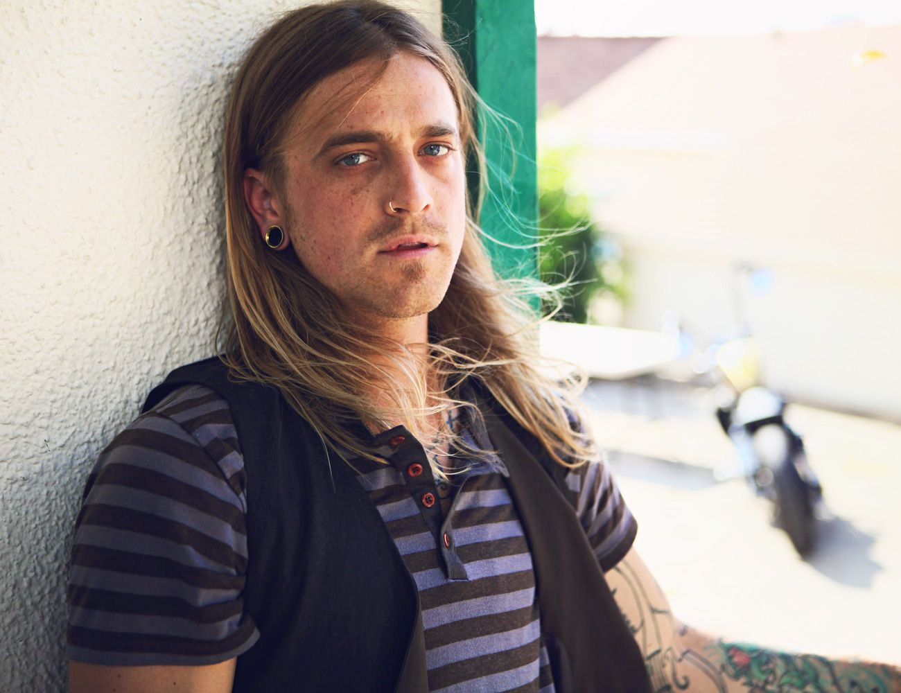 Person with long brown hair sits thoughtfully against a white wall in natural lighting.