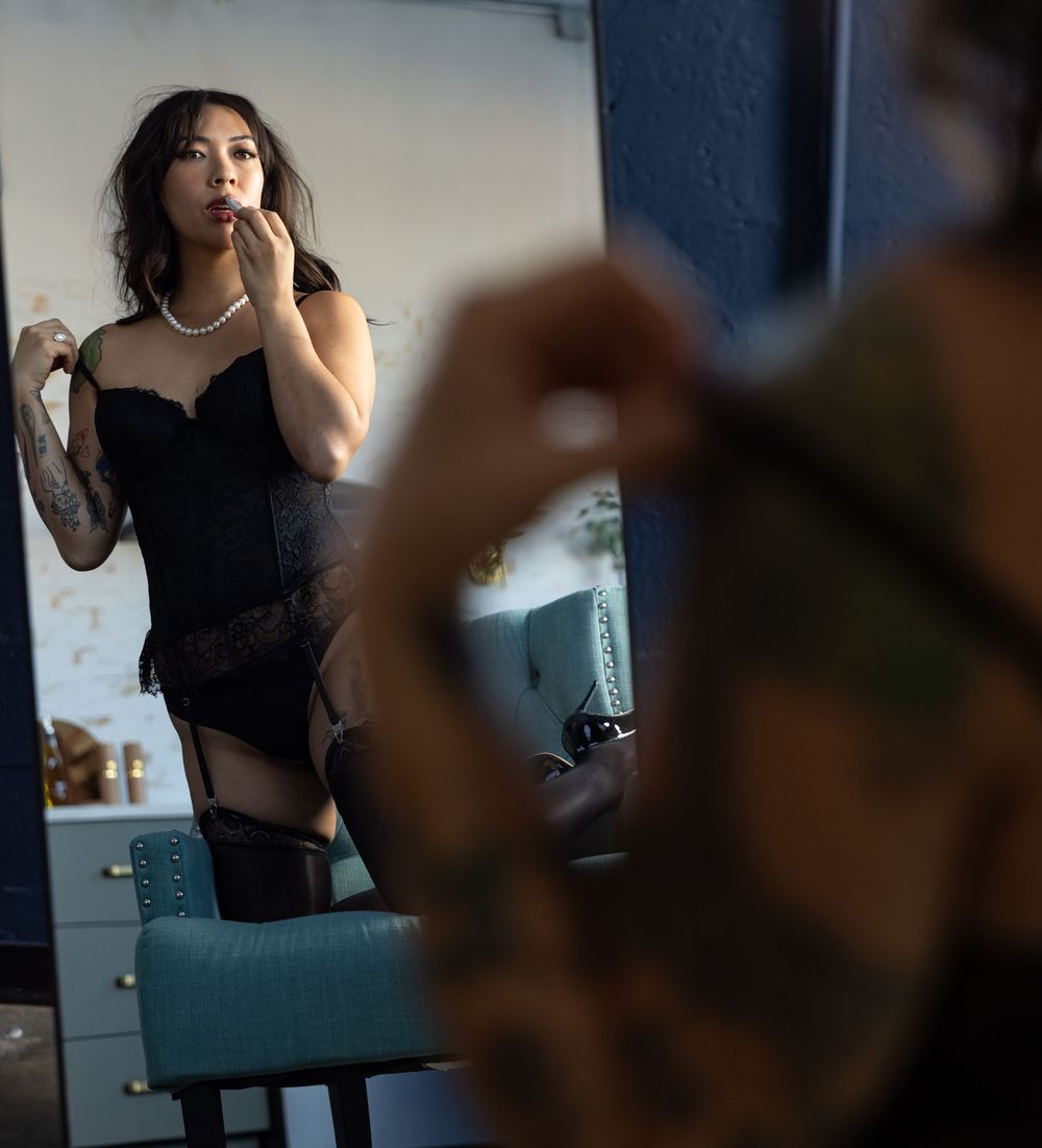 Woman in lingerie looking at her reflection as she puts on lipstick during boudoir and glamour photo shoot.