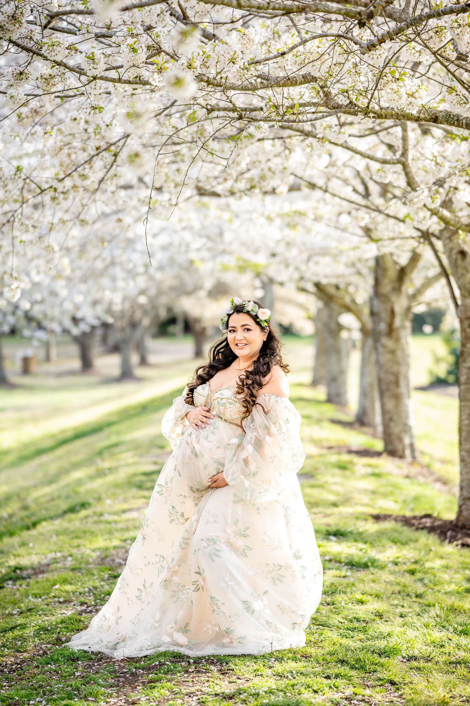 Expectant mother in a flowing lace dress poses under blooming cherry blossom trees in spring.