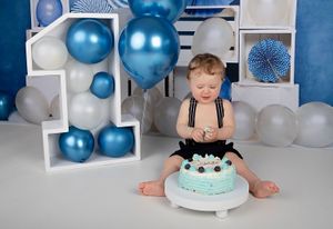 Baby sitting by turquoise birthday cake surrounded by blue and white balloons.