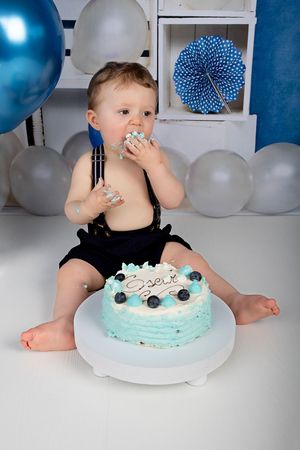 Baby in black suspenders eating cake during first birthday photoshoot with blue balloons.