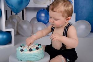 Baby in black suspenders reaching for decorations on light blue birthday cake.