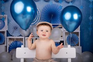 Baby in beige hat with blue balloon and paper fan decorations.
