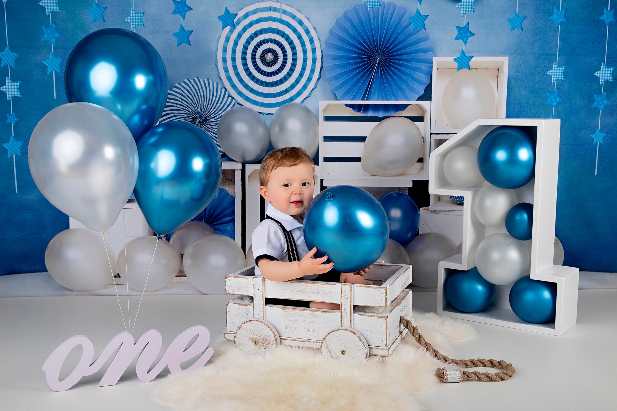 First birthday photo with baby in wooden cart prop surrounded by blue and white decorations.
