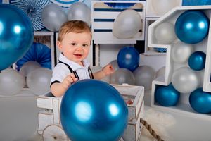 Baby in suspenders playing with blue balloon in decorated white and blue studio setting.