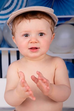 Baby wearing newsboy cap clapping hands against blue and white balloon backdrop.
