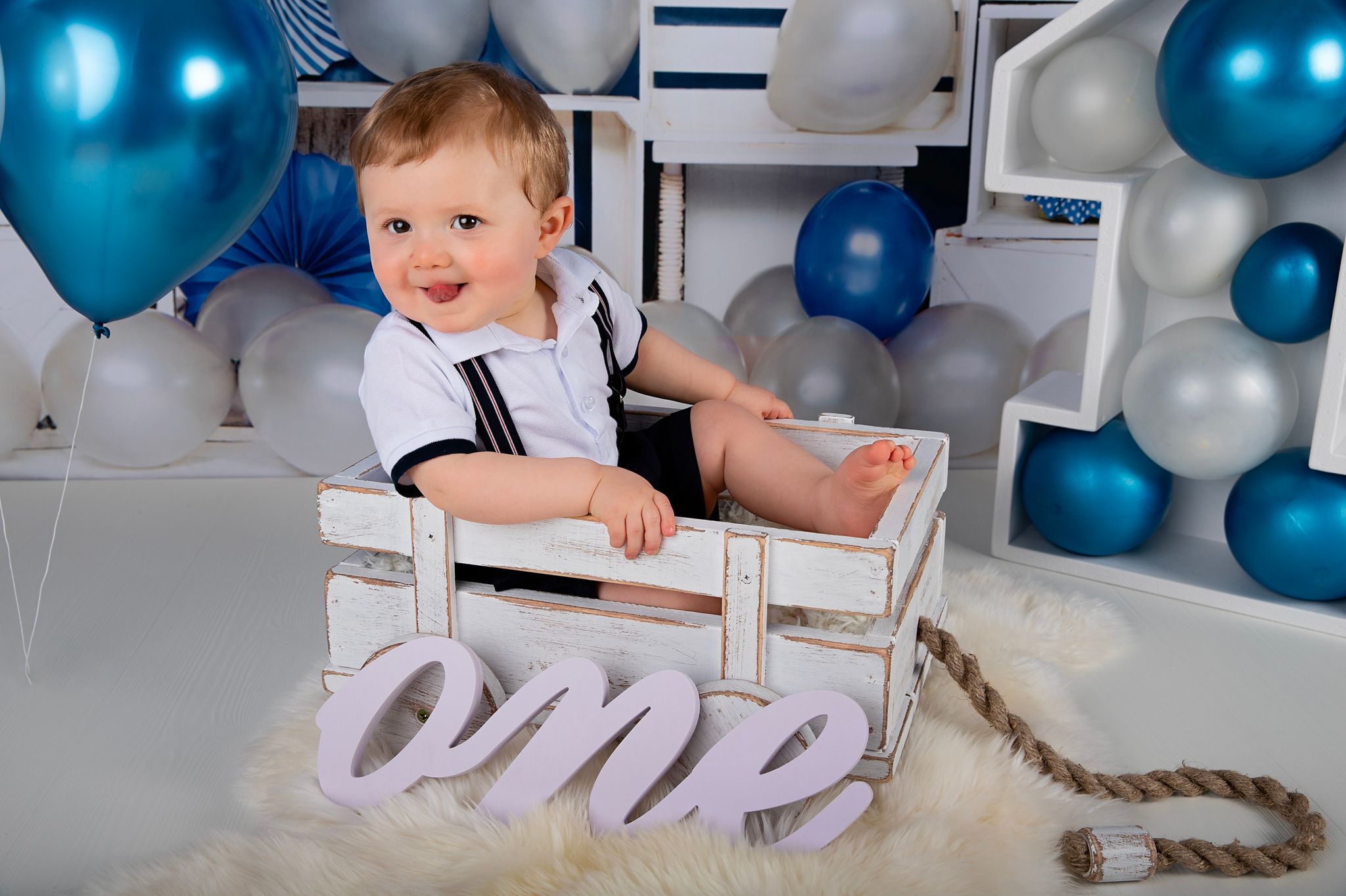 Smiling baby sitting in wooden cart with 'one' text decoration and balloon background.