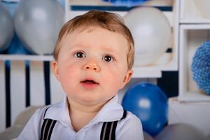 Portrait of baby in white suspenders against blue and white balloon backdrop.