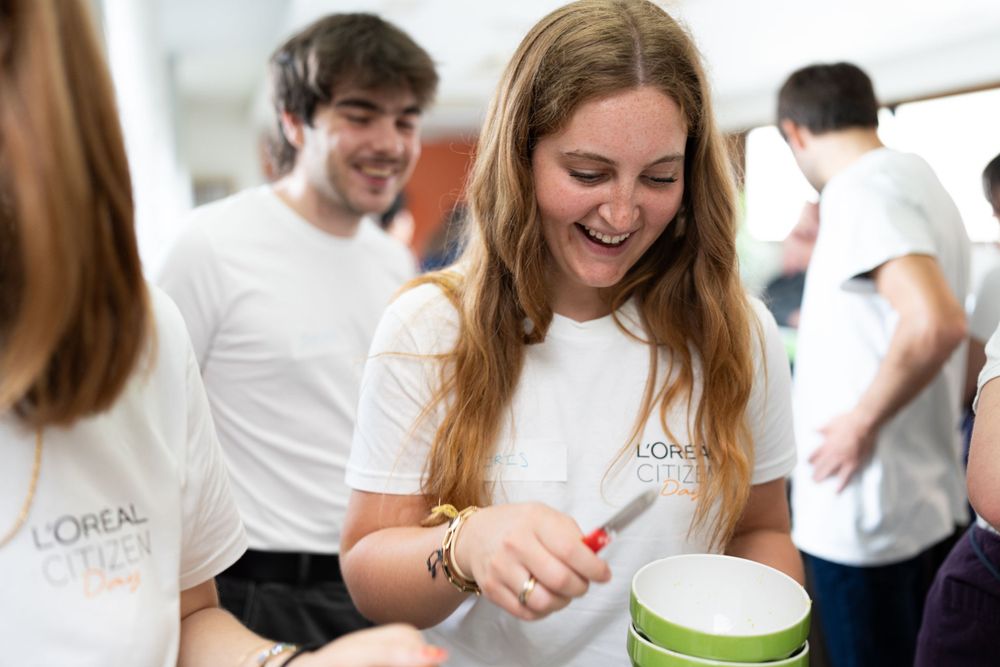 A young student in a white t-shirt smiles while looking down at a container during a social gathering.