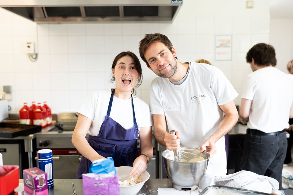 Two people in aprons work together while baking and mixing ingredients in a commercial kitchen setting.