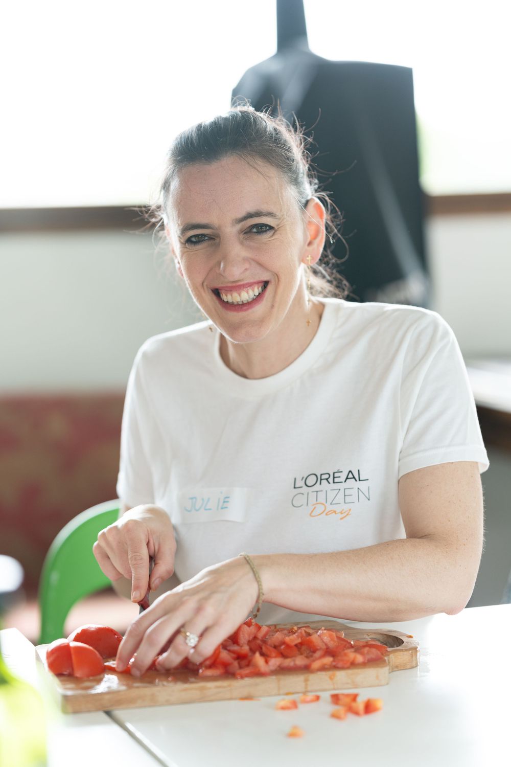 A person in a white L'Oreal t-shirt smiles while chopping fresh tomatoes on a wooden cutting board in a bright kitchen.