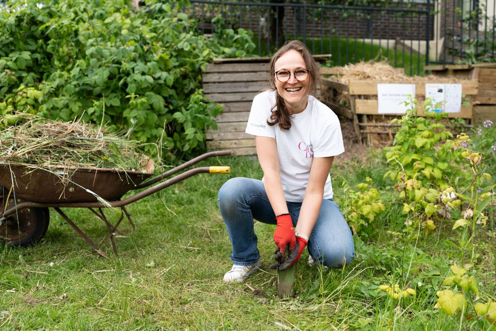 Gardener wearing red gloves kneels beside a rustic wheelbarrow in a lush backyard garden setting.