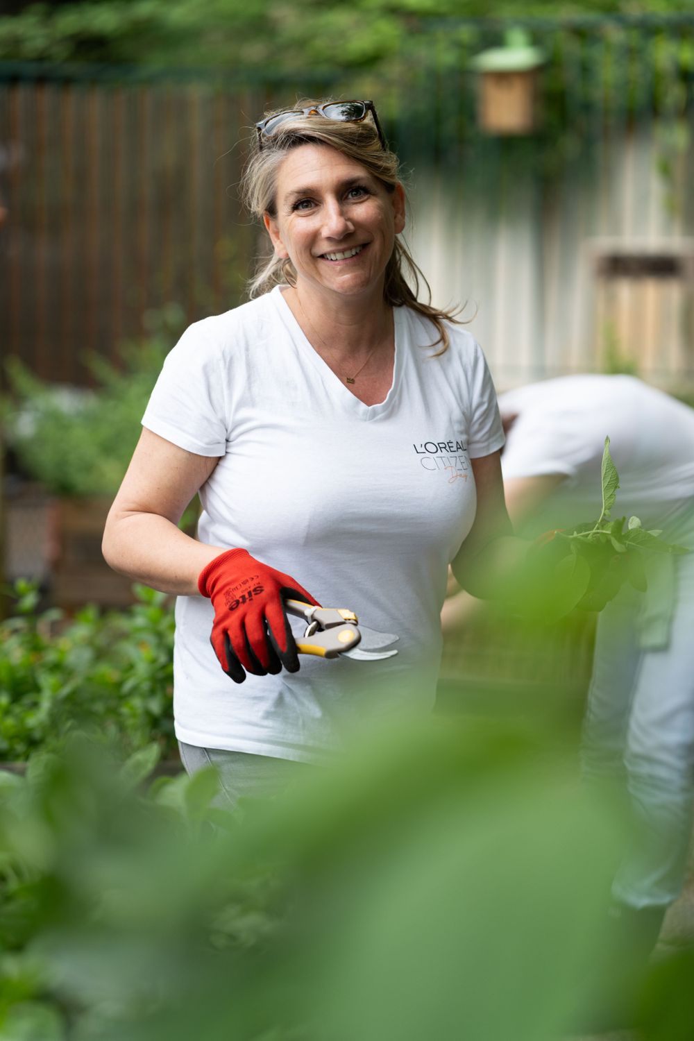 Gardener wearing white t-shirt and red gloves holds pruning shears in lush green garden setting.