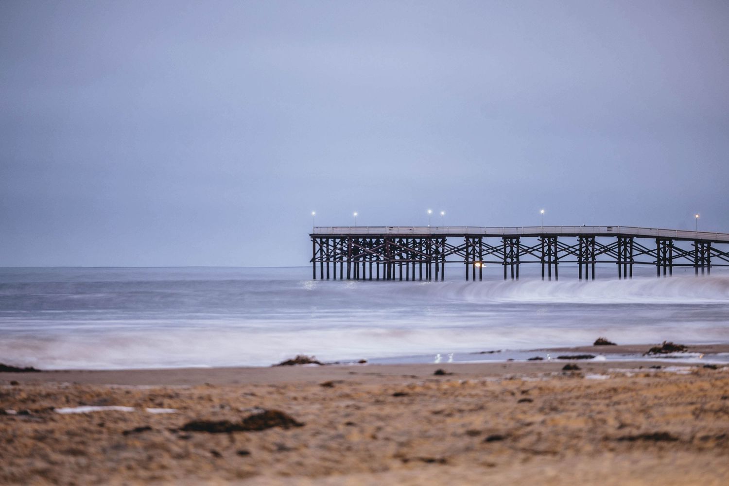 Long wooden pier stretches into a misty ocean with waves crashing on a sandy beach.