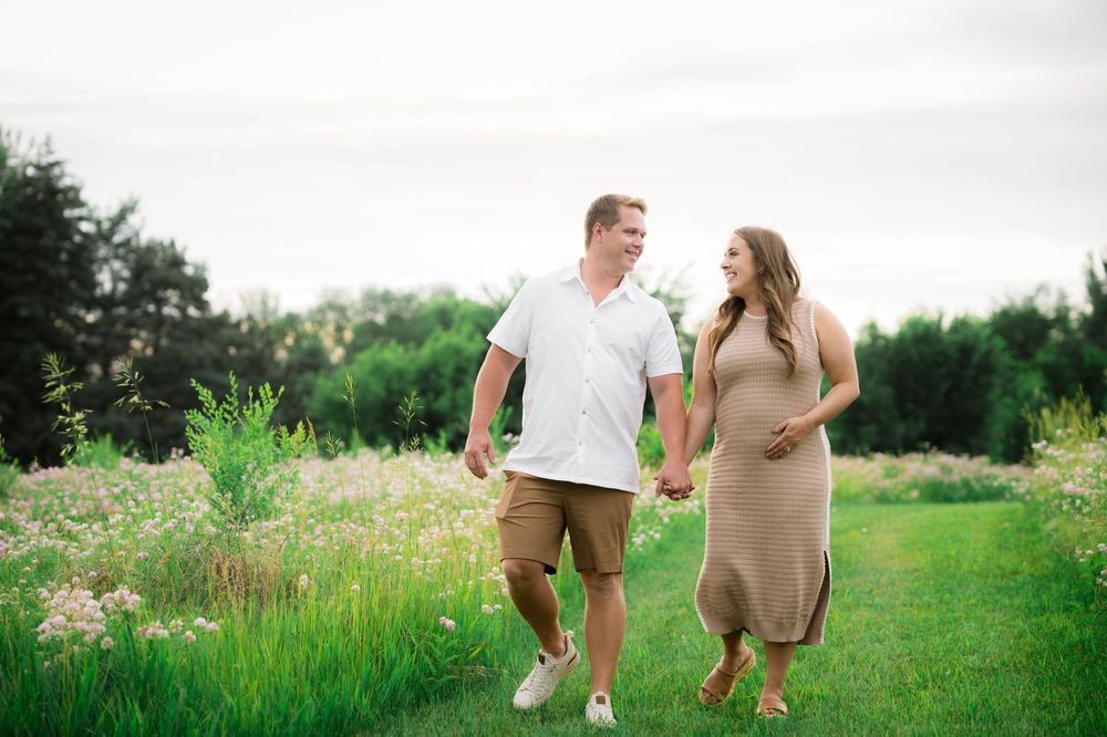 Happy expecting couple holding hands walking through green meadow with wildflowers during maternity photo session.