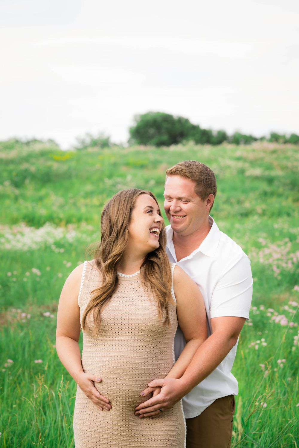 Smiling pregnant woman in beige dress with husband embracing her baby bump in lush green field.