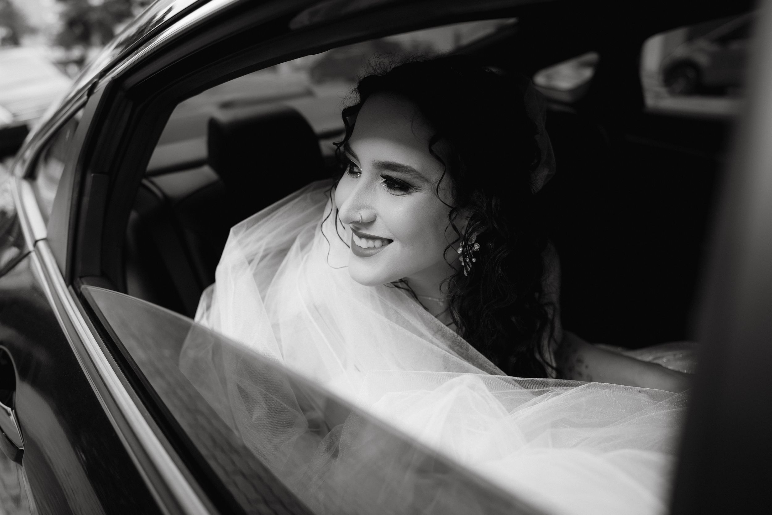 Black and white portrait of smiling passenger in wedding veil inside car.