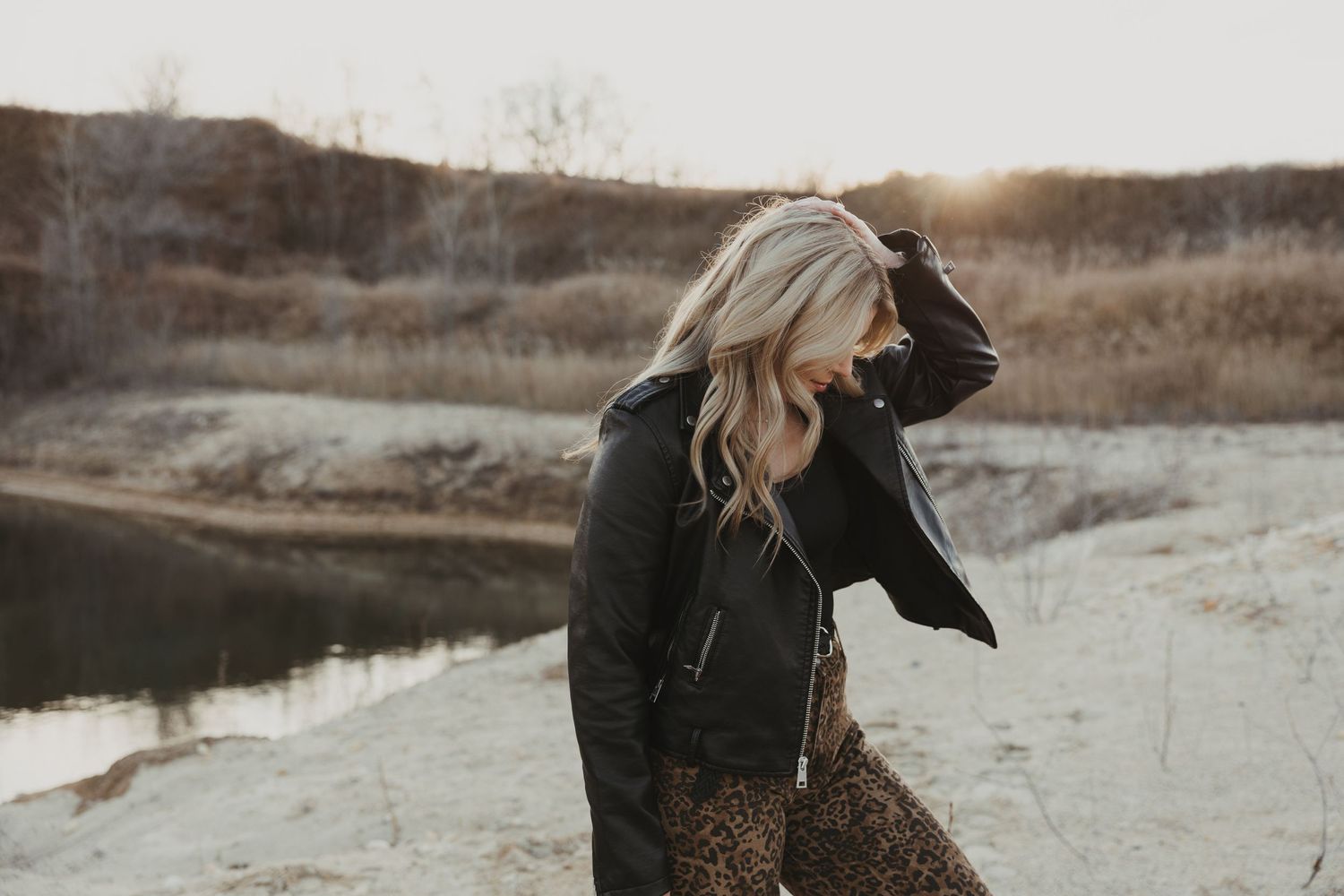 Blonde woman in leather jacket posing by a sandy riverside at sunset.