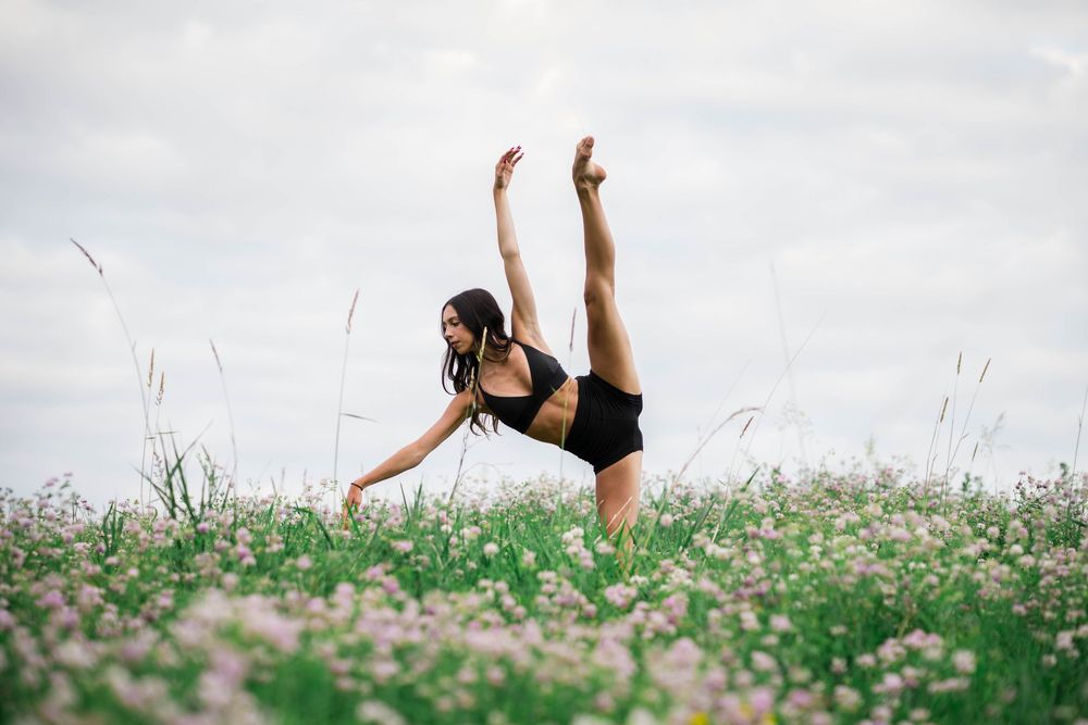 A dancer performs a graceful high kick in a field of wildflowers under a cloudy sky.