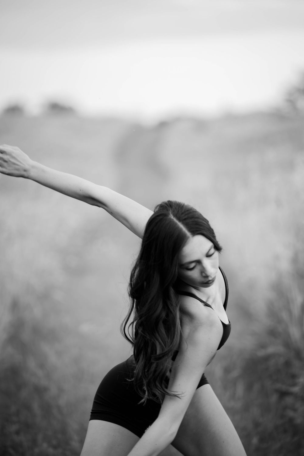 Black and white photo of a woman dancing gracefully in a field, arms extended, wearing a black bodysuit.