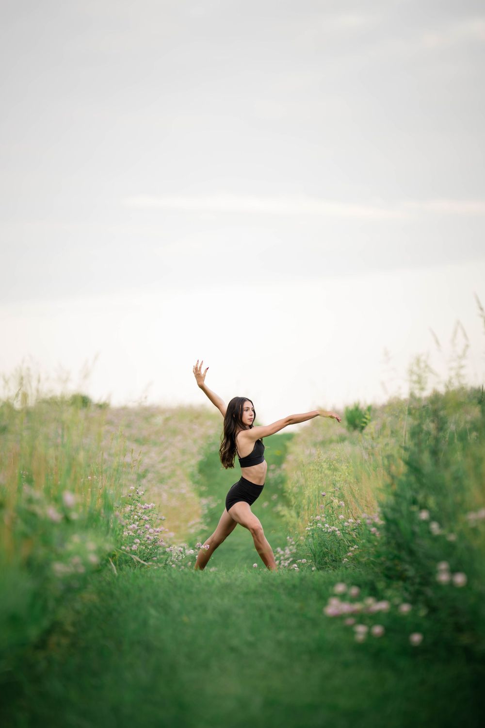 A graceful dancer in black activewear poses elegantly in a blooming green meadow under a soft cloudy sky.