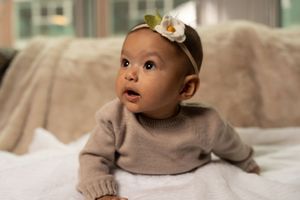 A baby wearing a white headband and beige sweater gazes upward while lying on a light-colored surface.