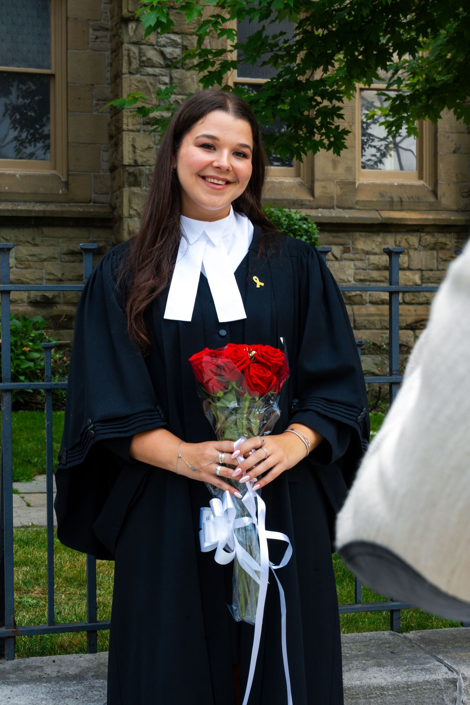 Graduate in black robe holds red roses while posing outdoors.