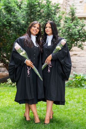 Graduates in black academic robes holding flowers pose on grass in front of stone building.