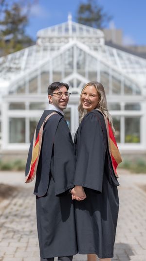 Two graduates in academic regalia pose together in front of a white greenhouse structure.