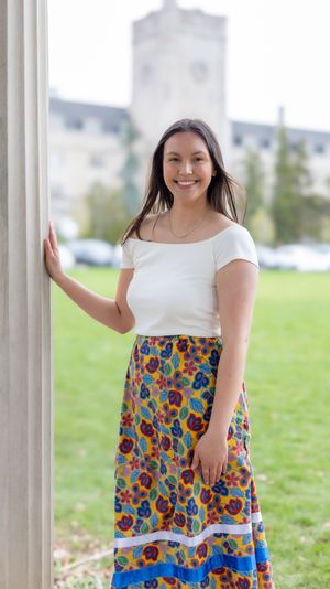 Standing by a column wearing a white top and colorful floral maxi skirt with a historic building in the background.