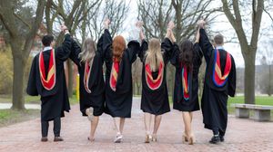Group of graduates in black robes and red stoles walking away on a tree-lined path.