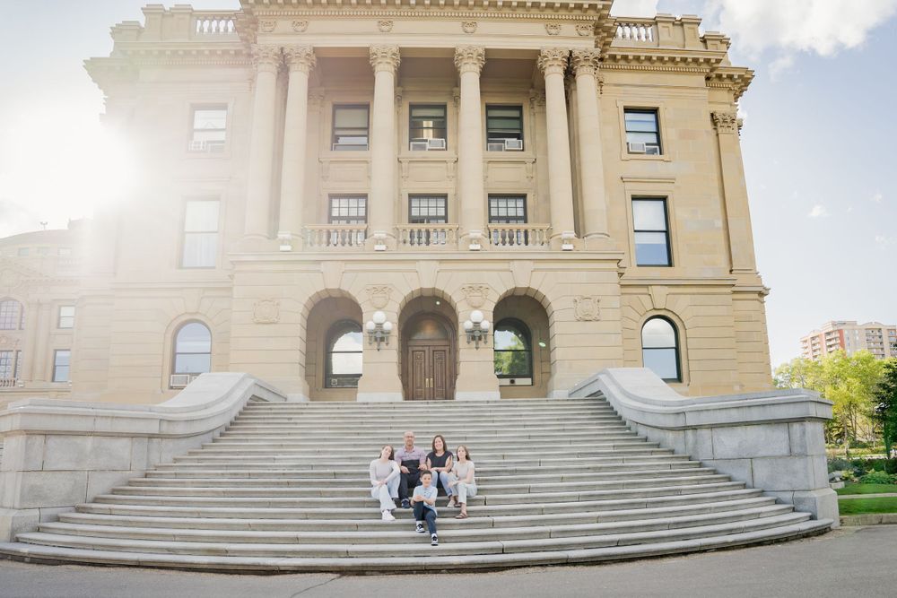 Wedding party stands on grand stone steps of historic building in bright sunlight.