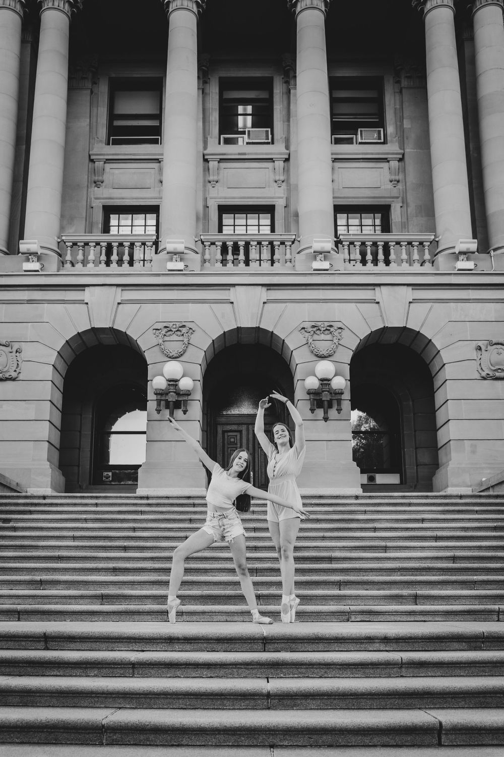 Two ballet dancers perform graceful poses on grand stone steps of a neoclassical building.