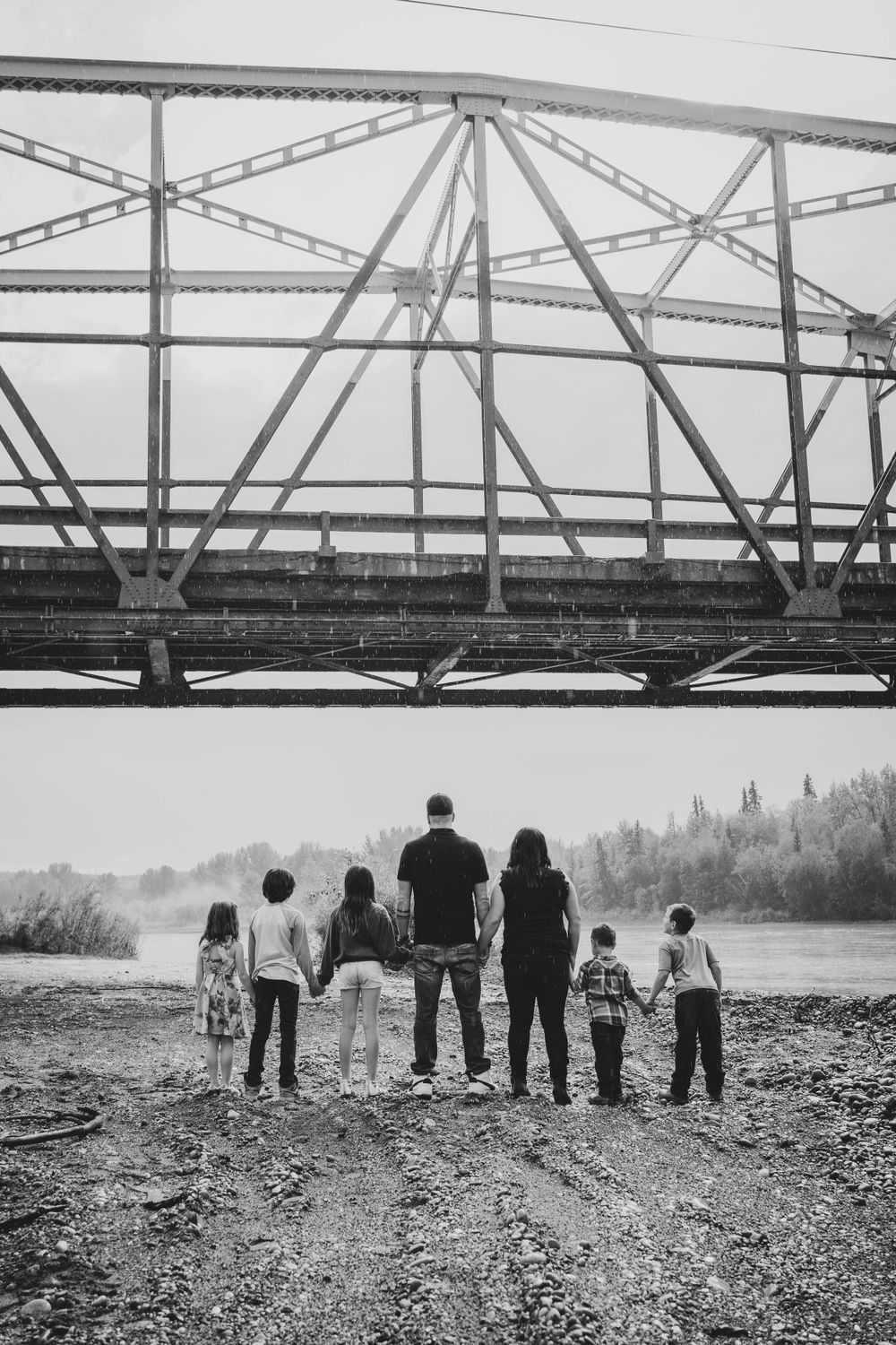 Family silhouette standing together under large metal bridge structure by riverside.