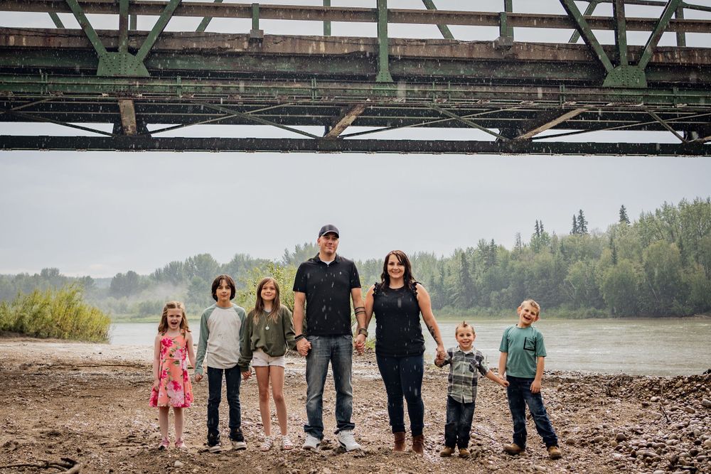 Family group photo beneath large metal bridge structure near river's edge.