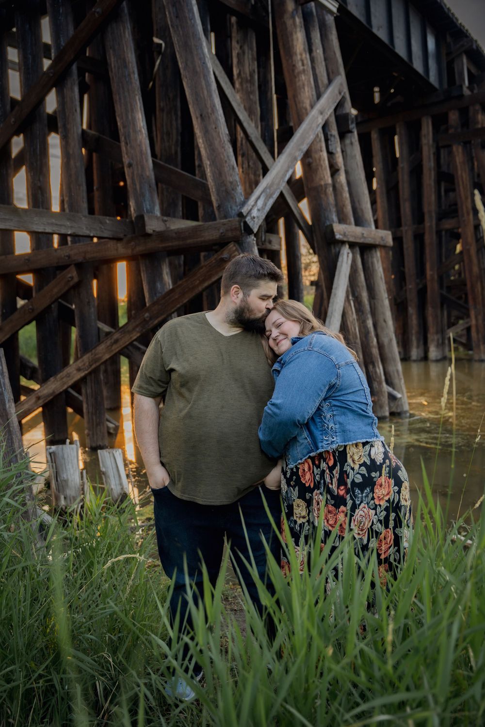 Couple shares intimate moment near wooden trestle bridge in tall grass.
