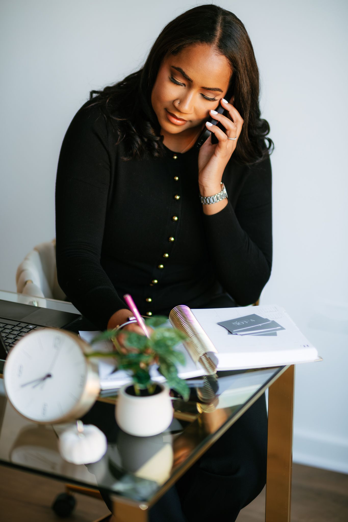 Woman in black cardigan working at desk while taking notes and talking on phone.