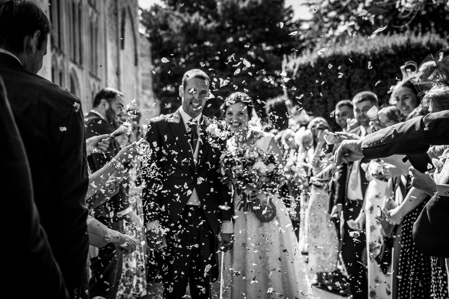Wedding guests throw confetti over a bride and groom during their outdoor celebration in black and white photography.