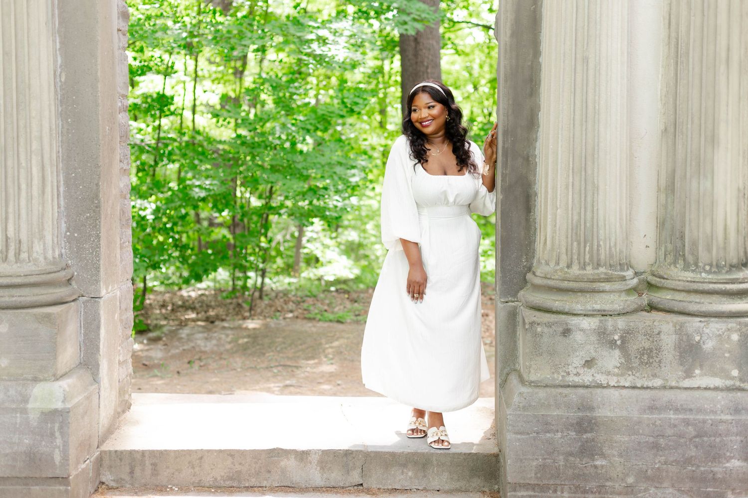 Romantic bridal photo featuring white dress among classical stone columns and green foliage.