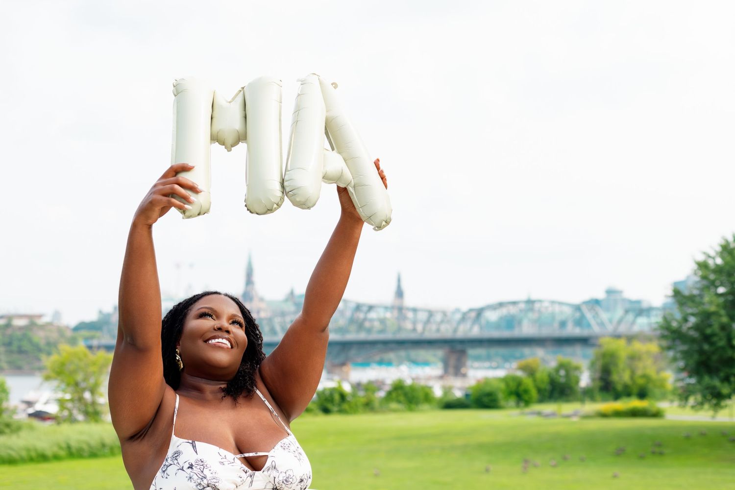 Happy woman in white dress holds up MHA letters outdoors near bridge.