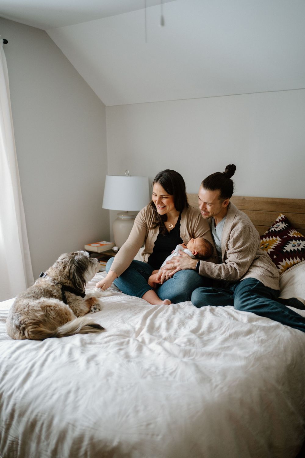 Couple sitting on bed with newborn baby boy and fluffy dog in cozy bedroom natural light from window-Muskoka Newborn photographer