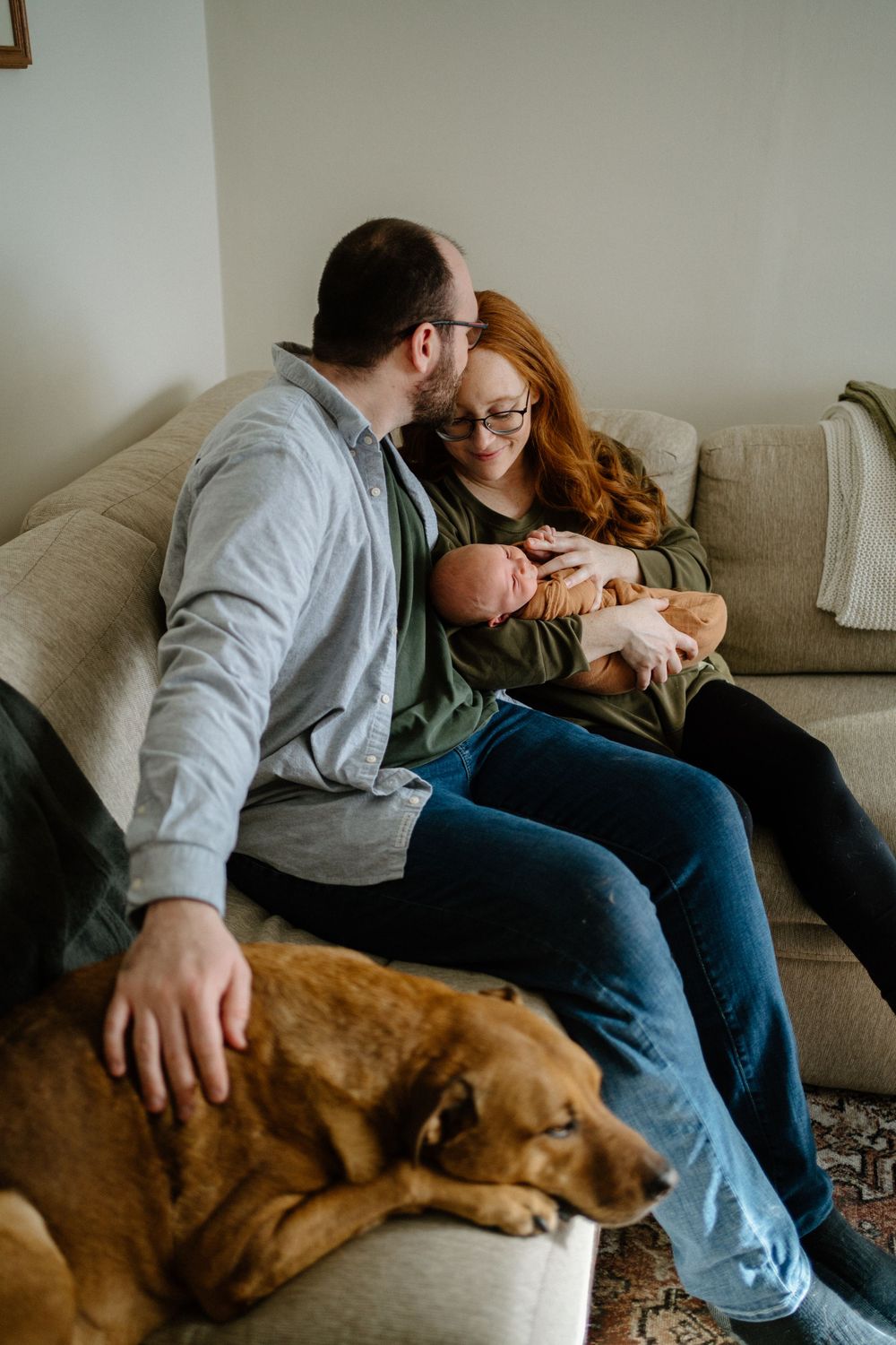 A couple sits on a grey couch with their newborn baby while their brown dog rests beside them-Muskoka Newborn photographer