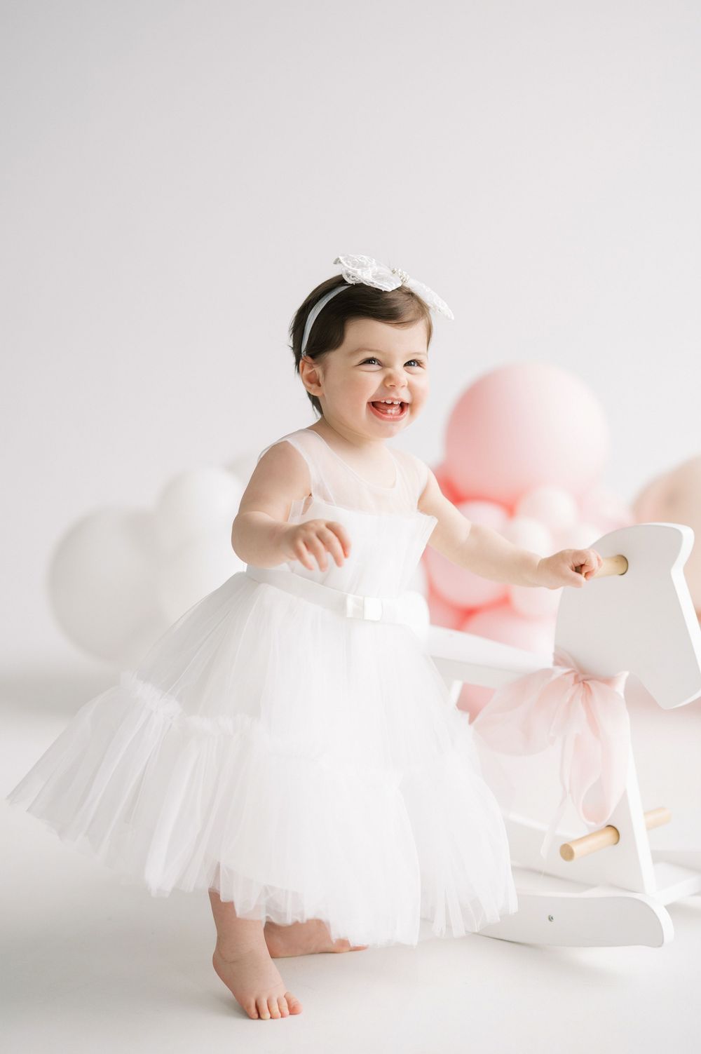 A small child wearing a white tulle dress laughs joyfully next to pink balloons and a white rocking horse.
