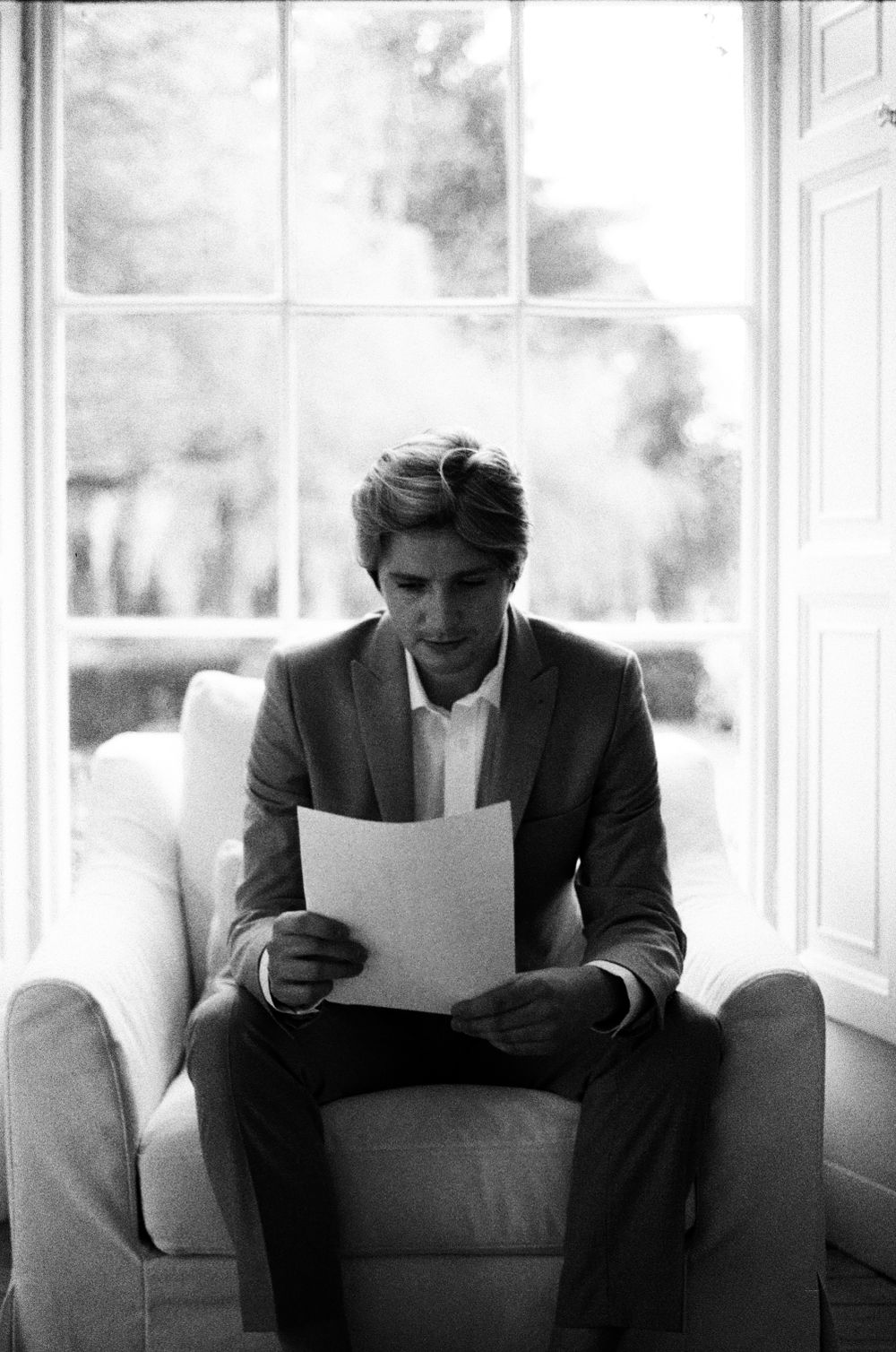 Black and white portrait of a groom reading a letter by a window.