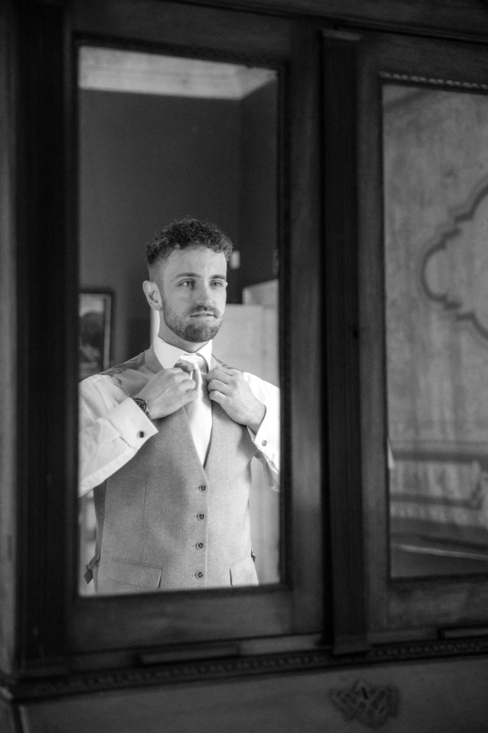 Groom adjusting bowtie in mirror reflection, wearing light colored vest and white shirt.