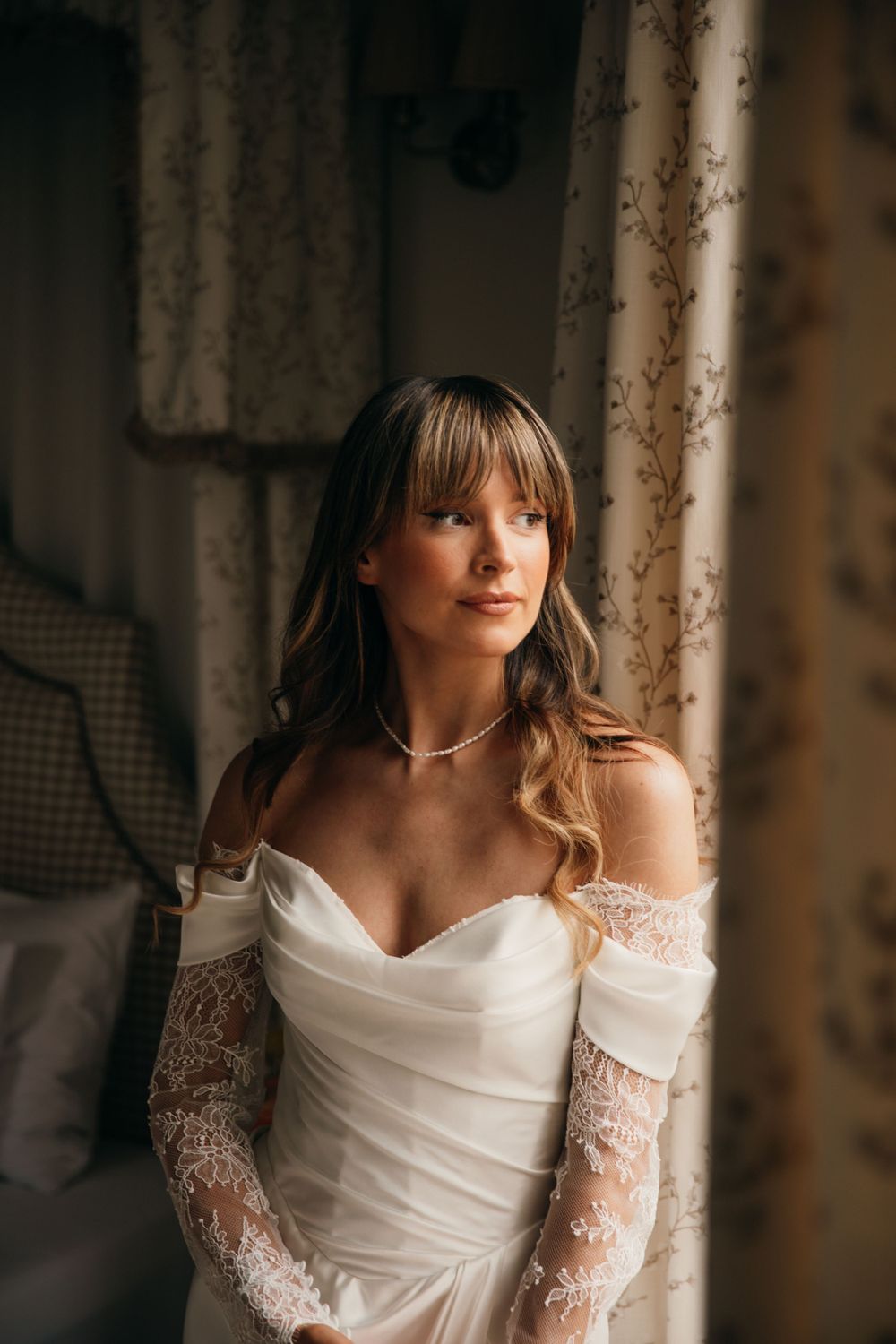 Bride in off-shoulder lace wedding dress poses by window with natural light.