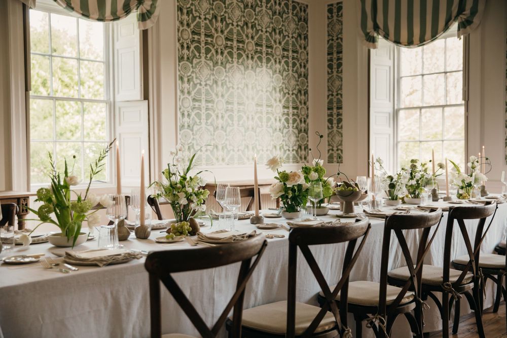 Elegant dining table set with white linens, cross-back chairs, and floral centerpieces near bright windows.
