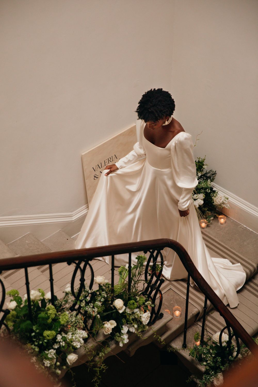Bride in elegant white gown with long sleeves walking down decorated staircase with floral arrangements.